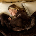 A blonde woman rests on white pillows, wrapped snugly in a large, soft Snuggle Luxe Blanket, looking cozy and content.