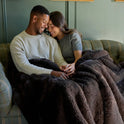 A couple smiles and holds hands on a couch, wrapped together in the cozy Snuggle Luxe Blanket.