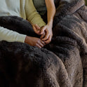 Two people sit close together under the Snuggle Luxe Blanket, holding hands for comfort and warmth. Only their hands, arms, and parts of their torsos are visible beneath the thick, dark brown plush fabric.