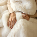 A person drapes themselves in the Snuggle Luxe Blanket, their hands resting on its soft, plush fabric, while wearing a beige sweater.