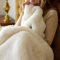 A woman sits on a chair, wrapped in a Snuggle Luxe Blanket in Vanilla, holding the plush blanket close to her face. She wears a long-sleeve beige top, with a softly glowing gold lamp in the background.