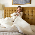 A woman sits on a bed with a mustard tufted headboard, smiling as she wraps herself in the plush Snuggle Luxe Blanket. A painting above her completes the cozy atmosphere.
