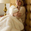 A woman smiles on a bed with a tufted headboard, wrapped in the Snuggle Luxe Blanket. A gold lamp sits on the bedside table in the background.