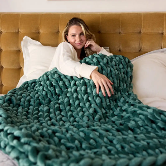 A woman relaxes in bed, leaning against white pillows and a tan tufted headboard, wrapped in the cozy comfort of an ultra-soft Emerald Velvet Weighted Blanket.