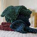 A velvet weighted blanket in green, navy blue, and burgundy hues is stacked on a bed with a white quilted cover for cozy comfort. A blurred lamp and headboard appear in the background.