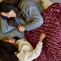 A woman and a young girl lie side by side on a couch, smiling and wrapped together in a Velvet Weighted Blanket. The woman wears a gray sweater, while the girl is dressed in a cream top.
