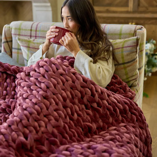 A woman relaxes on a plaid armchair, wrapped in the Ruby Velvet Weighted Blanket for cozy comfort, holding a red mug near her face and looking content.