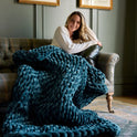 A woman with long hair sits on a sofa, smiling, wrapped in a sapphire Velvet Weighted Blanket. Framed pictures on the wall and a patterned rug enhance the cozy comfort of the room.