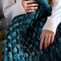 Close-up of a person’s hands holding the Velvet Weighted Blanket in teal. They wear a cream sweater and sit on a plaid couch, enjoying the ultra-soft, chunky comfort.
