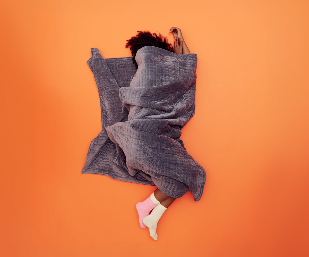 A person lies on an orange surface, wrapped in a large, quilted gray Gravity Original Weighted Blanket (10–25 lb) filled with glass beads.