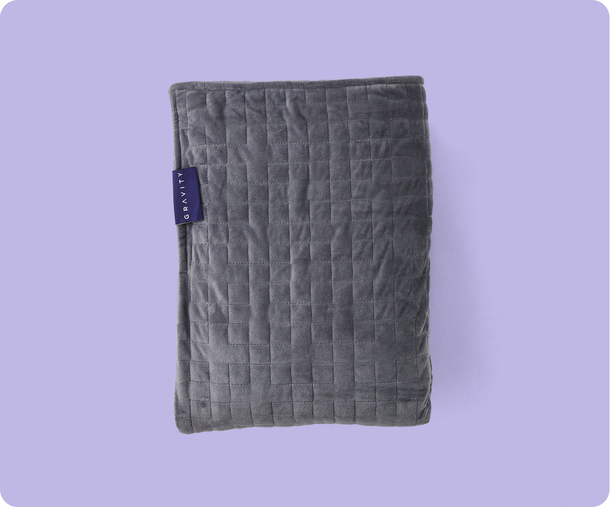 A folded dark gray Gravity Original Weighted Blanket (10–25 lb) with a breathable cover, a stitched grid pattern, and a small rectangular brand tag on the side.