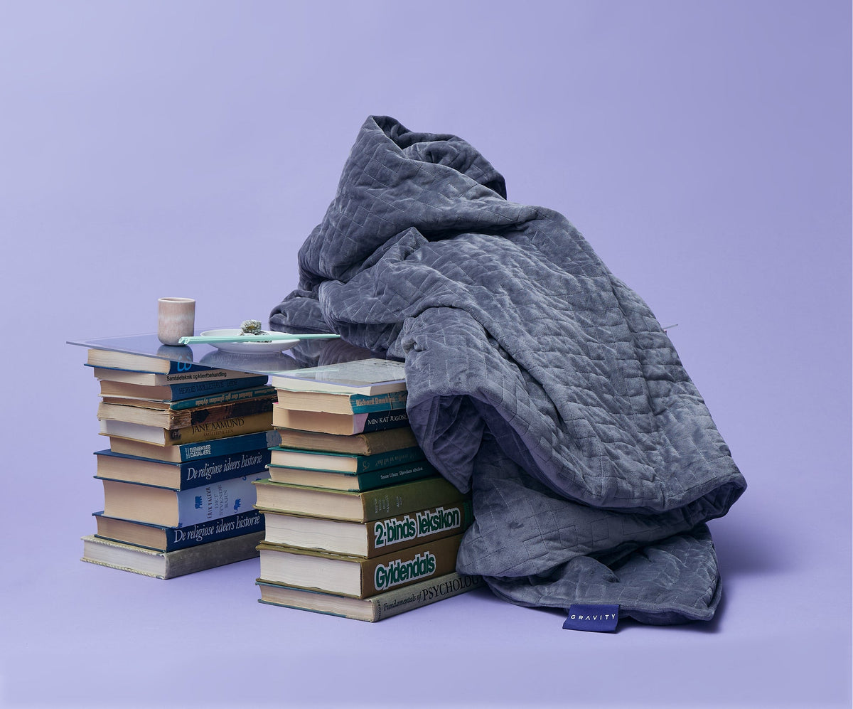 A gray quilted Gravity Original Weighted Blanket (10–25 lb) with a breathable cover draped over a glass coffee table supported by two stacks of books.