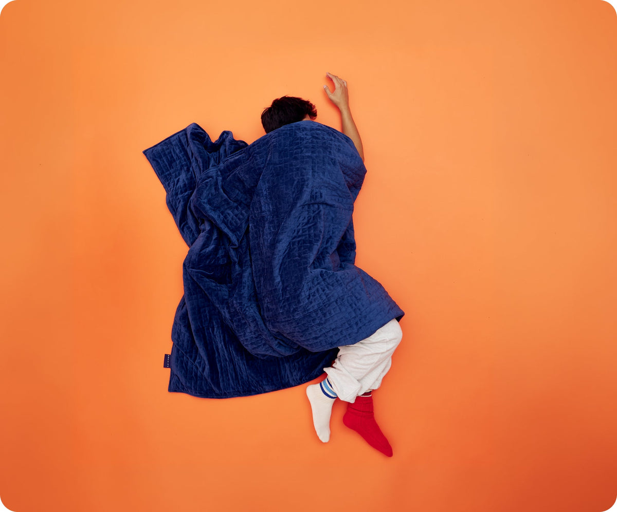 A person sleeping on an orange surface under a dark blue quilted Gravity Original Weighted Blanket (10–25 lb) with a breathable cover.