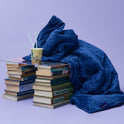 A blue quilted Gravity Original Weighted Blanket (10–25 lb) draped over two stacks of books, with a cup of instant noodles on a clear acrylic tray balanced between them, the breathable cover resting atop the stacks.