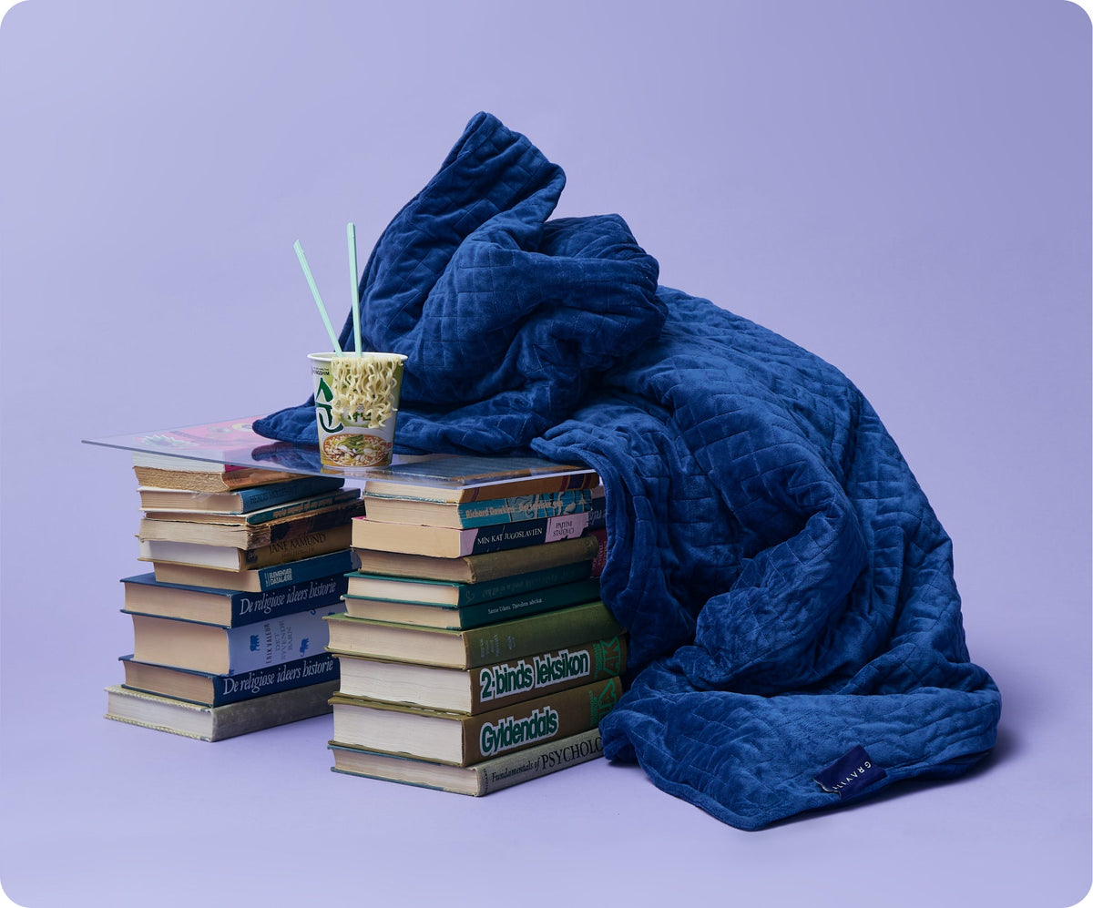A blue quilted Gravity Original Weighted Blanket (10–25 lb) draped over two stacks of books, with a cup of instant noodles on a clear acrylic tray balanced between them, the breathable cover resting atop the stacks.