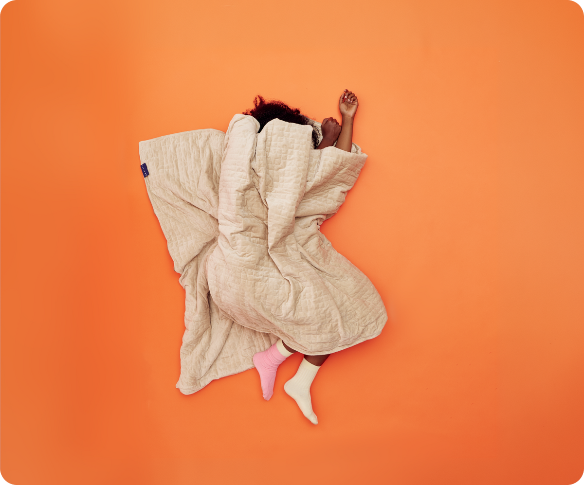A person sleeping on an orange surface, covered by a Gravity Original Weighted Blanket (10–25 lb) filled with glass beads.