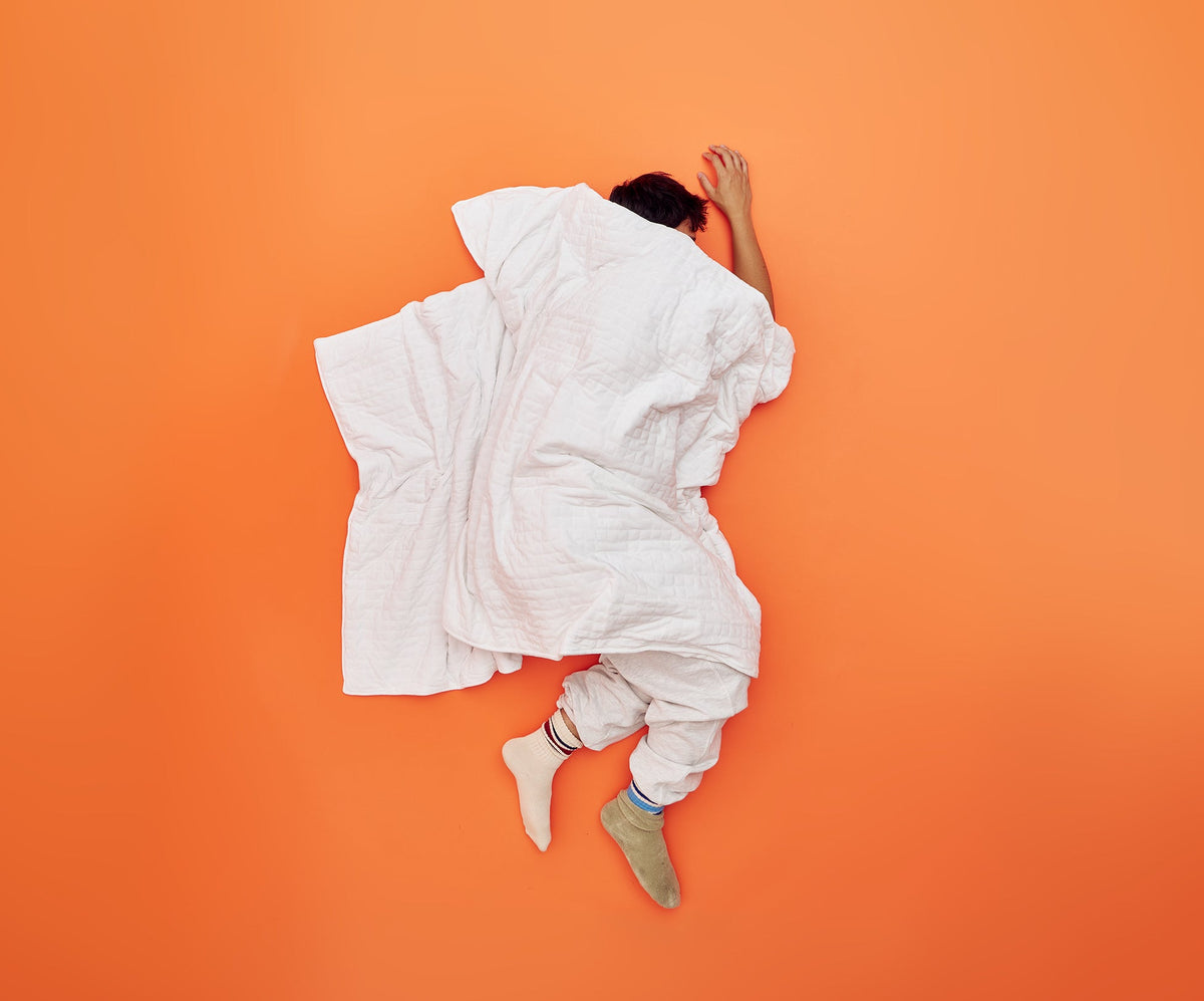 A person sleeping on an orange surface while covered by a white, quilted Gravity Original Weighted Blanket.