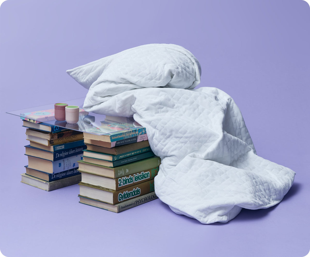 A white quilted Gravity Original Weighted Blanket (10–25 lb) with a breathable cover draped over a glass-topped table made of two stacks of books.