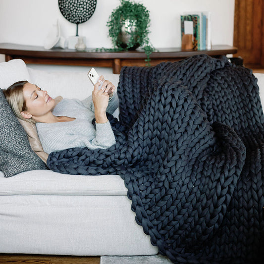 A woman relaxes on a couch, wrapped in a Knit Weighted Blanket and smiling at her phone. Decorative pillows, plants, and a wooden console with ornaments create a cozy atmosphere in the background.