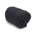 The Knit Weighted Blanket, featuring a chunky hand-knitted black braided design, is neatly rolled up on a plain white background.