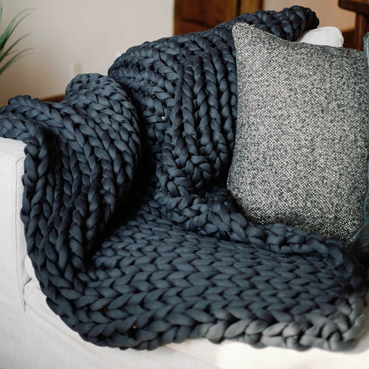 A Knit Weighted Blanket in dark blue is draped over a light-colored sofa beside a textured gray and white pillow.