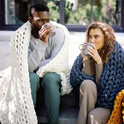 A man and a woman sit on outdoor steps, wrapped in Knit Weighted Blankets and sipping from mugs. They look relaxed and cozy, with greenery and large windows in the background.