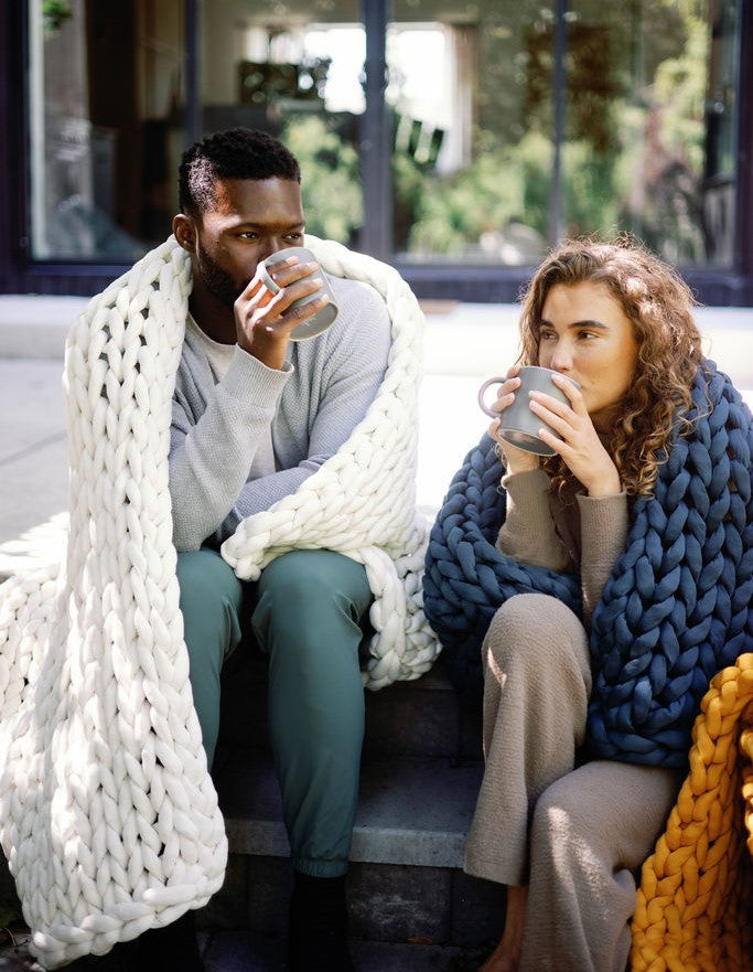 A man and a woman sit on outdoor steps, wrapped in Knit Weighted Blankets and sipping from mugs. They look relaxed and cozy, with greenery and large windows in the background.