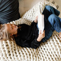 A child relaxes on the Knit Weighted Blanket, resting their head on an adult’s lap while reading, dressed in a black sweater and jeans; only part of the adult in a dark shirt is visible.
