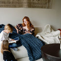 A red-haired woman smiles on a sofa with a Knit Weighted Blanket, holding a mug. Two children sit nearby—one reading—in a cozy living room with warm décor.