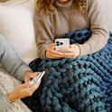 Two people sit on a bed covered with chunky knit blankets, including the Knit Weighted Blanket in blue and white. Each holds a smartphone—one wears a tan sweater, both focused on their screens.