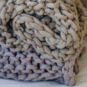A close-up of the Knit Weighted Blanket, rolled up on a flat surface. Its thick, light gray yarn gives a cozy, textured look thats perfect for comfort and relaxation.