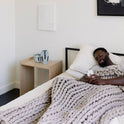 A man relaxes in bed under the Knit Weighted Blanket, browsing his phone. Beside him, a small wooden table holds two ceramic mugs and a vase in a simply decorated room.