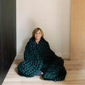 A smiling child sits on a light wooden floor, wrapped in a dark green Knit Weighted Blanket with chunky knit, against a light-colored wall featuring black and wood paneling.