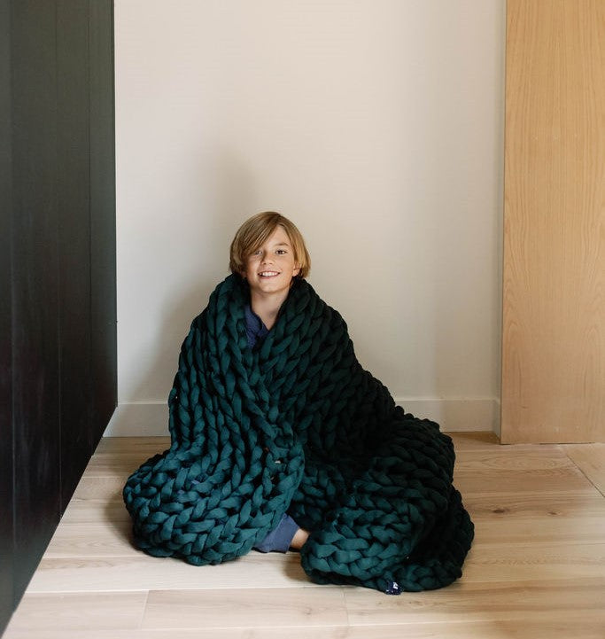 A smiling child sits on a light wooden floor, wrapped in a dark green Knit Weighted Blanket with chunky knit, against a light-colored wall featuring black and wood paneling.