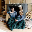 Two people sit on the floor by a fireplace, wrapped in a Knit Weighted Blanket. One smiles while sipping from a mug. Firewood and a furry stocking add warmth to the cozy scene.
