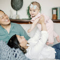 A smiling woman lifts a happy baby girl in a pink outfit and blue bow headband while they relax under the super soft Knit Weighted Blanket; a man in a green shirt sits beside them on the couch.