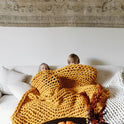 Two children are cuddled on a white sofa under the Knit Weighted Blanket. A table with a pumpkin and dried flowers sits in front, while a patterned rug decorates the wall behind them.
