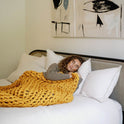 A woman with curly hair relaxes on a white bed, draped in the Knit Weighted Blanket in chunky yellow. Abstract art decorates the wall above.