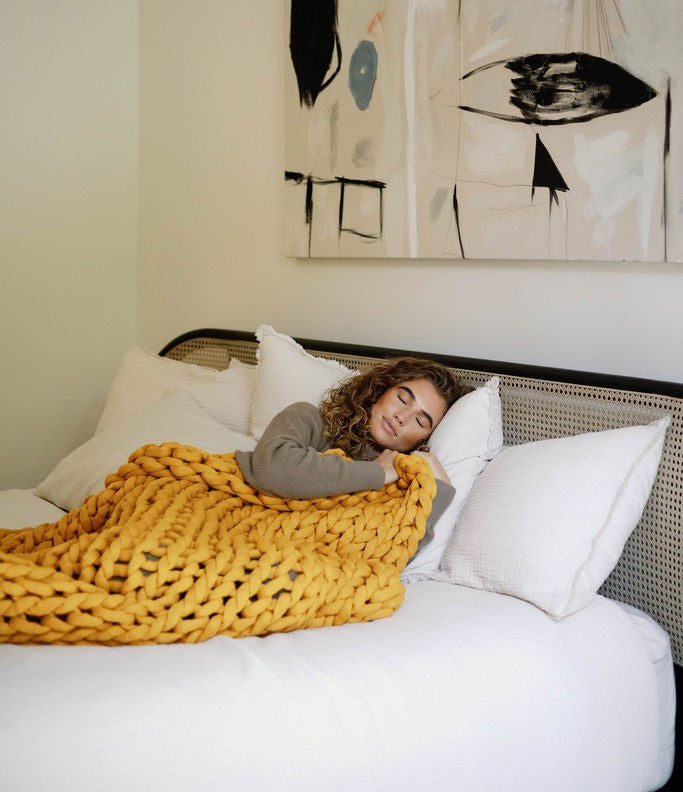 A woman with curly hair relaxes on a white bed, draped in the Knit Weighted Blanket in chunky yellow. Abstract art decorates the wall above.