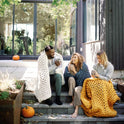 Three people sit on outdoor steps wrapped in Knit Weighted Blankets, smiling and sipping hot drinks. Pumpkins are around them, with a modern house featuring large windows in the background.
