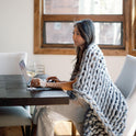 Wrapped in a Knit Weighted Blanket, a person sits at a table using a laptop and holding a glass of white wine in a bright room with modern furniture and a large window.