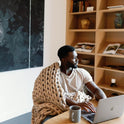 A man wrapped in a Knit Weighted Blanket sits at a table with a laptop and mug, looking to the side. Shelves with books and decor are behind him, along with a large abstract painting on the wall.