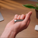 A hand flips a sleek ONO Roller Steel fidget accessory with the thumb. In the background, a blurred plant, part of a laptop, and a wireless earbud case rest on a wooden desk.