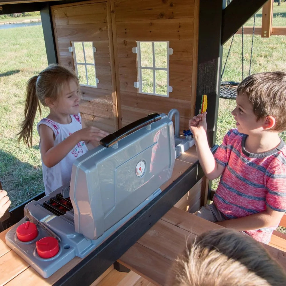 Two children enjoy imaginative play with a toy grill inside the Playhouse Backyard Discovery Canyon Creek Cedar Swing Set, as sunlight streams in. The girl pretends to cook while the boy holds a toy food item in this fun outdoor playground.