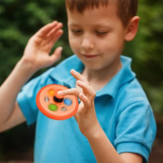 A young boy in a blue shirt from Mainland China smiles and focuses as he spins the Pop Fidget Toy Gyro—a spring mechanism bubble pop rubber tire—on his finger, with a blurred green outdoor background.