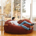 A man in a white shirt and jeans relaxes on the Fuf XXL Foam Filled Bean Bag Chair Deep Terracotta 6 Feet in a bright room with large windows, gazing outside at trees and a water view.