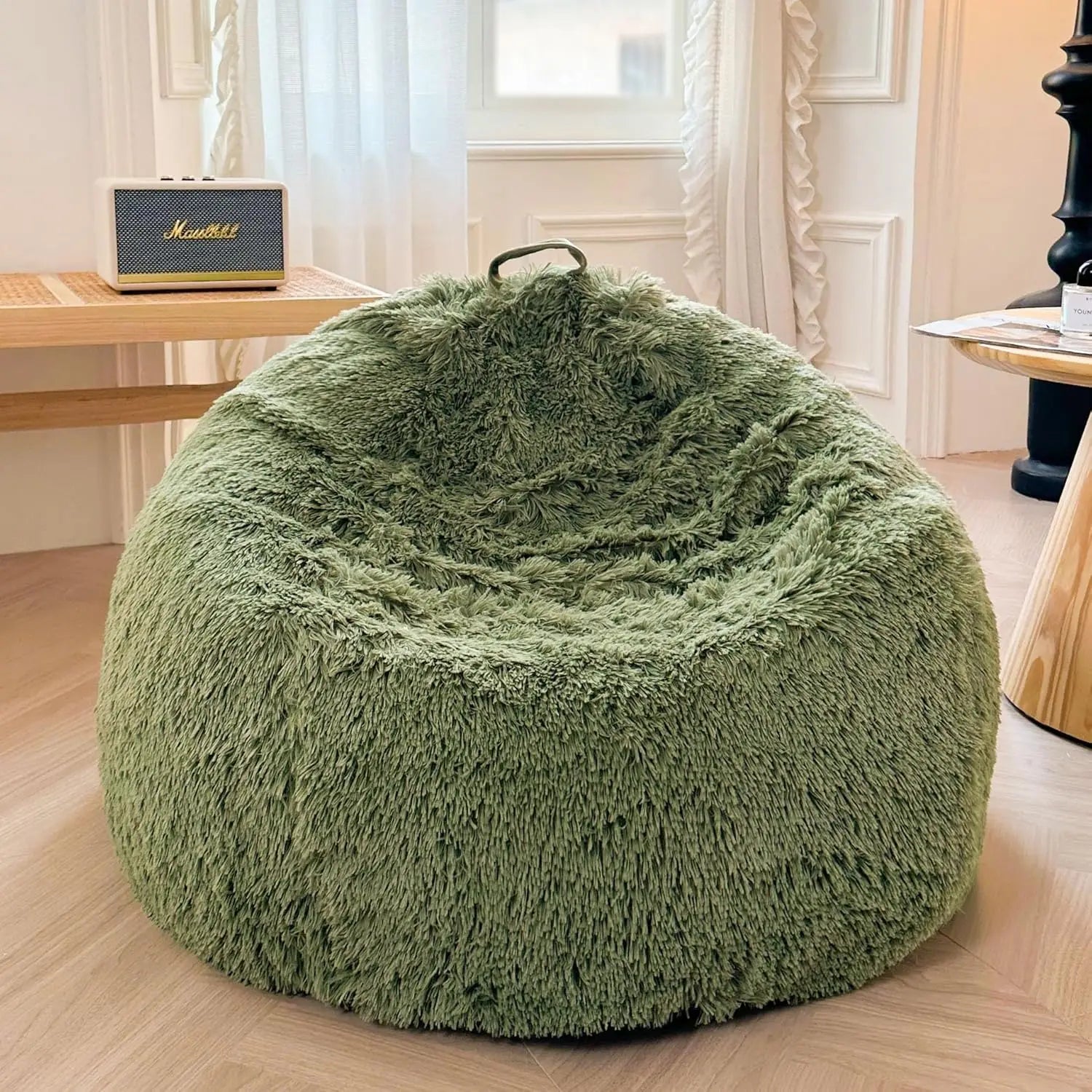 A sage green Faux Fur Comfy Bean Bag Cover (No Filler Storage Chair) sits on a wooden floor in a cozy room with light walls and curtains. Nearby tables hold a radio, while natural light from the window highlights the plush seating.