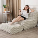 A woman relaxes barefoot on the Standard-Green Bean Bag Sofa Chair with Footstool, wearing a white t-shirt and black shorts, holding a remote. A side table and plant are nearby as she lounges comfortably in this modern reading chair for adults.