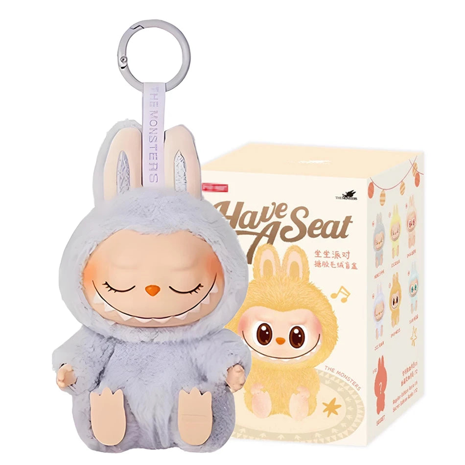 The Cute Labubu The Monsters Box Toy Sweetheart Vinyl Doll in Style B with box features a grinning bunny plush keychain with closed eyes and floppy ears, plus a decorative box showing the matching Labubu character and Have A Seat text.