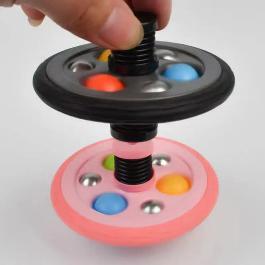 A hand holds the Pop Fidget Toy Gyro, a two-tiered fidget with black and pink layers, colorful balls, metal spheres, and a spring mechanism—made of quality materials from Mainland China.